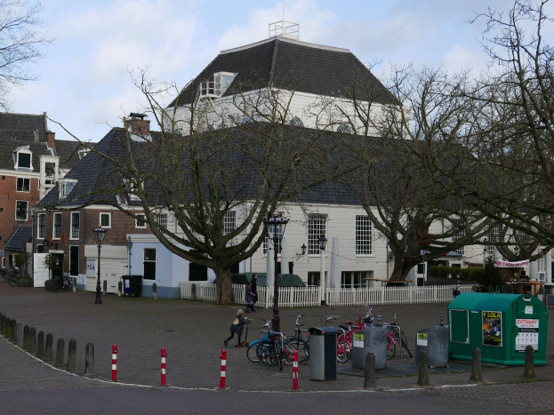 Eine Gruppe von Fahrrädern, die vor einem Rathausesgebäude geparkt sind, mit Menschen in der Nähe, umgeben von Bäumen, Pfählen, einem Zaun, Gebäuden und einem bewölkten Himmel.