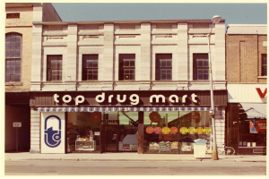 Ein Drogengeschäft an einer Stra√enecke mit einem Gebäude, einem Schild, einem Stra√enpfahl, einem abgestellten Fahrrad, einem Müllcontainer und einem bewölktem Himmel.