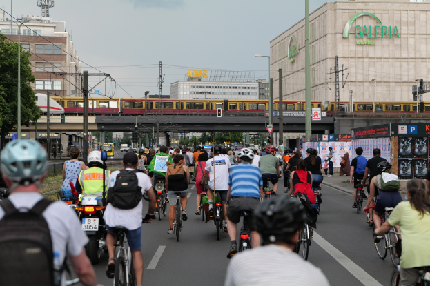 Eine Gruppe von Menschen, die auf Fahrrädern eine Straße entlangfährt, die von hohen Gebäuden gesäumt ist, einige tragen Helme und Taschen, mit einem Zug auf einem Bahngleis, Strommasten, Bäumen und einem klaren blauen Himmel im Hintergrund.