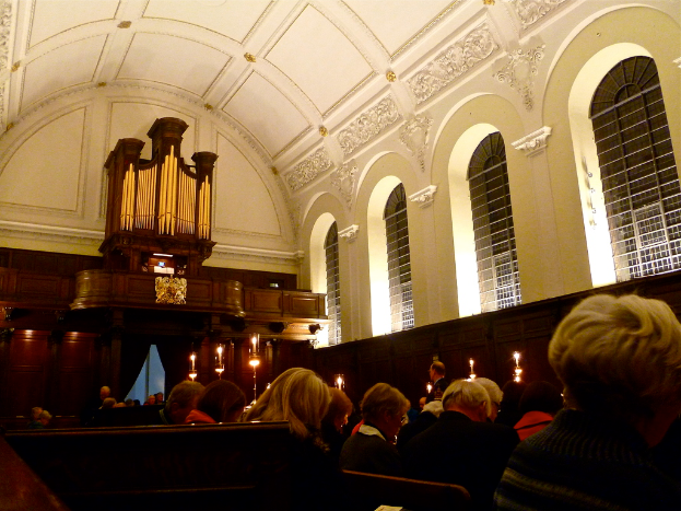 Der Innenraum einer Kirche mit sitzenden Menschen und Kerzen davor, mit weißen Wänden und Decke.
