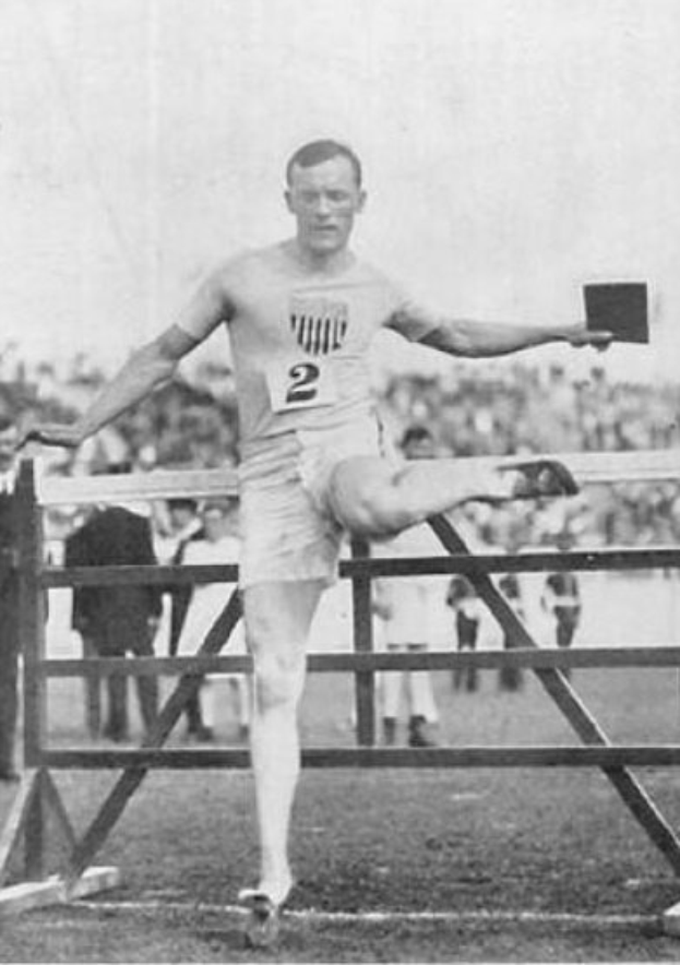 Ein Schwarz-Weiß-Foto eines Mannes in Sportkleidung, der über ein Hindernis springt, mit Zuschauern im Hintergrund und dem Himmel oben.