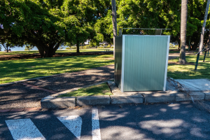 Eine mobile Toilette am Straßenrand in einem Park, umgeben von Bäumen und Gras, mit einem Wasserlauf und einem klaren blauen Himmel im Hintergrund.