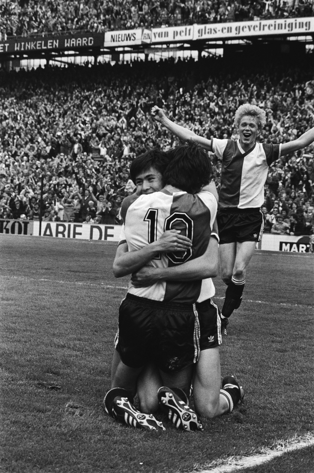 Drei Fussballspieler feiern ein Tor, indem sie sich auf dem Boden umarmen, vor einem jubelnden Publikum in einer schwarz-weissen Stadionszene.