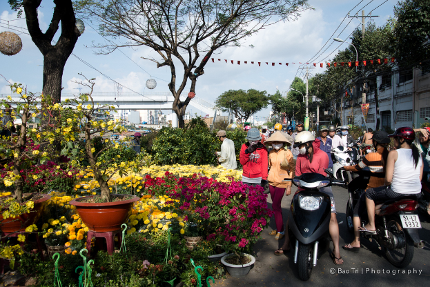 Eine Gruppe von Menschen in Helmen und Mützen, einige gehen und andere fahren mit Fahrrädern auf einer Straße, mit Pflanzen, einem Blumentopf auf einem Hocker, Bäumen, einem Pfahl, Drähten, einer Lampe, einer Brücke und Gebäuden in der Nähe.