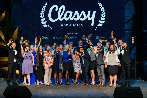 Eine Gruppe von Menschen auf einer Bühne, die lächeln und Auszeichnungen halten, mit Lautsprechern unten und einem Hintergrundbild mit der Aufschrift "Classy Awards 2019."