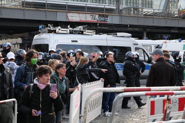Eine Gruppe von Menschen steht vor Polizeifahrzeugen hinter Barrieren, einige tragen Helme und halten Telefone, mit einer Brücke und Gebäuden im Hintergrund während einer Demonstration in Berlin.