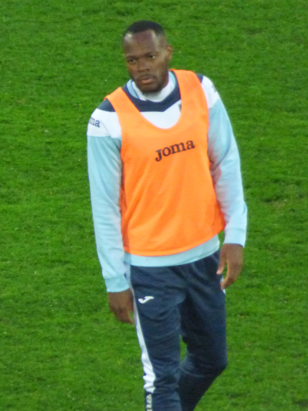 Ein Mann in einem orangen T-Shirt und blauen Hosen, der wie ein Fussballspieler aussieht, geht entschlossen über ein saftig grünes Feld.
