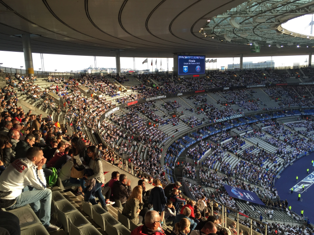 Große Zuschauermenge im Allianz Arena Stadion bei einem Fußballspiel, mit einer Bühne rechts, Fahnen, Stangen, einem Bildschirm und sichtbarem Himmel im Hintergrund.