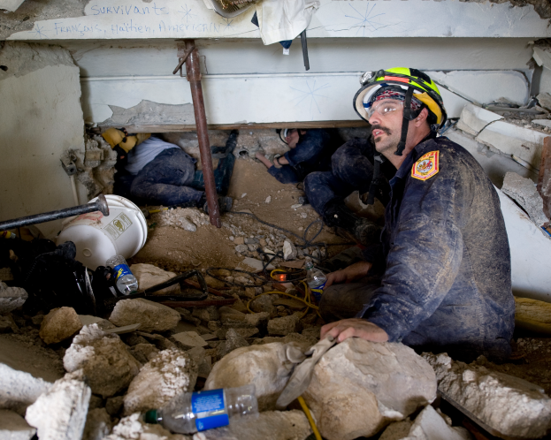 Ein Feuerwehrmann in einem Helm sitzt zwischen Trümmern, darunter Steine und Flaschen, mit einer Wand im Hintergrund, die einen Text trägt, an der Stelle eines israelischen Luftangriffs.