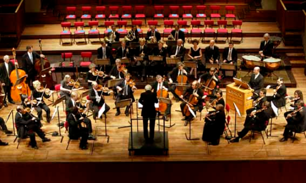 Ein großes Orchester auf einer Bühne mit einem Dirigenten davor, der verschiedene Instrumente spielt, mit Ständern und anderen Gegenständen drumherum und Stühlen und Treppen im Hintergrund.