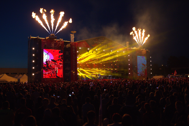 Eine große Menschenmenge auf einem Musikfestival mit einer Bühne, auf der Musiker unter hellen Scheinwerfern spielen, umgeben von Bannern, Zelten links und einem Feuerwerk, das den dunklen Himmel erhellt.