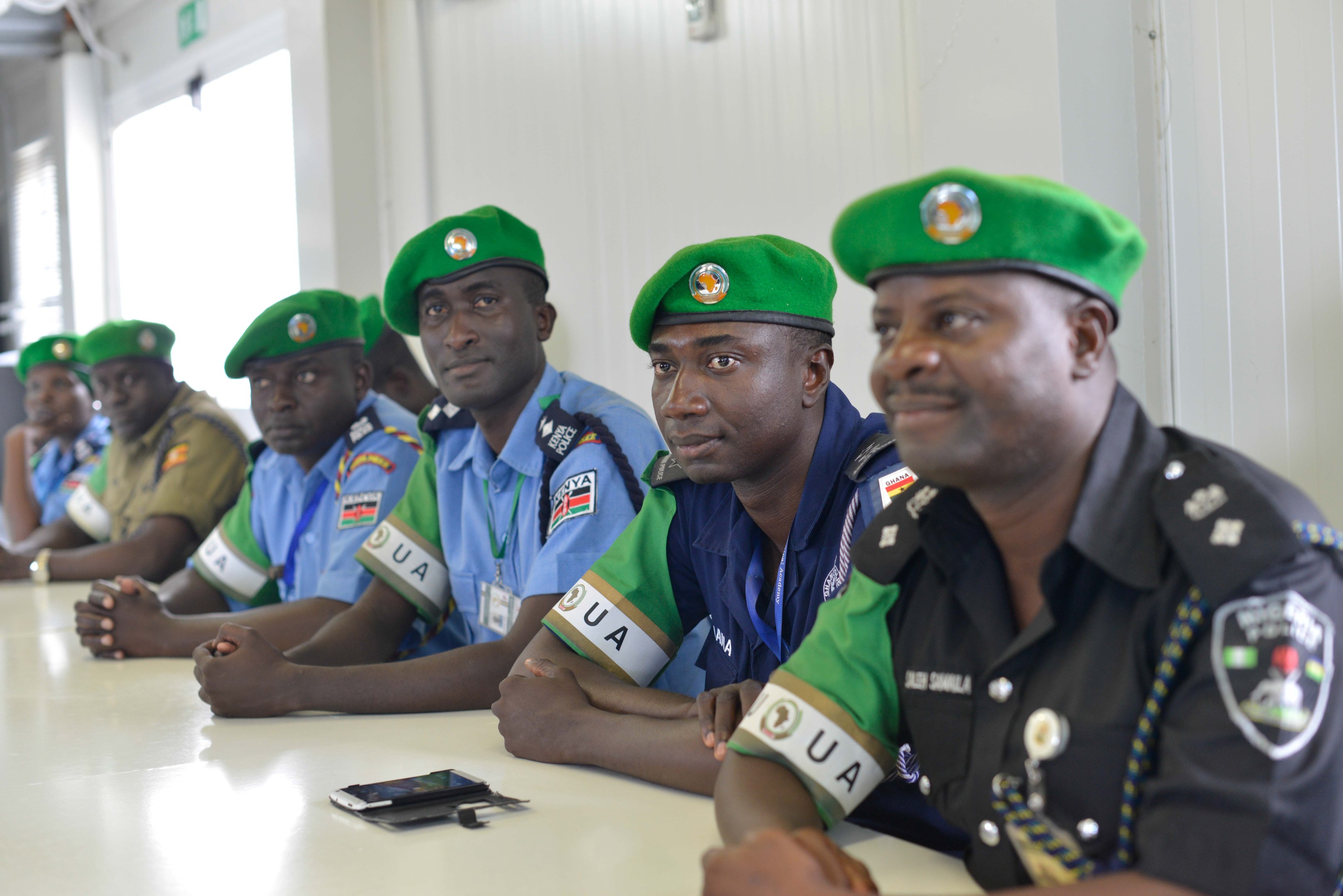 Eine Gruppe von Polizeibeamten sitzt an einem Tisch während einer Schulung, mit Mobiltelefonen und anderen Gegenständen auf dem Tisch, in einem Raum mit Fenstern und Deckenleuchten.