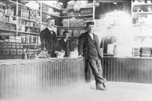 Drei Männer stehen an einem Tresen in einem Laden, umgeben von verschiedenen Gegenständen, mit einem Regal voller Bücher und anderen Gegenständen im Hintergrund, während eines Raubüberfalls.