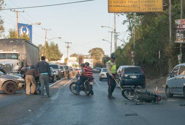 Gruppe von Menschen um ein verunglücktes Motorrad auf dem Seitenstreifen mit mehreren Fahrzeugen, darunter ein Lastwagen, im Hintergrund und B├Ąumen, Masten, Lichtern, Schildern und Himmel.