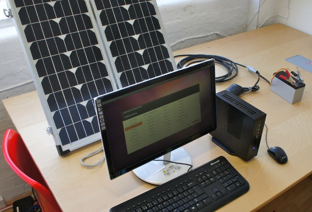 Ein Computersystem mit Tastatur, Maus und CPU auf einem Tisch neben einem Solarpanel, einer Batterie, Chips und Kabeln, mit einem roten Stuhl und einer weißen Wand im Hintergrund.