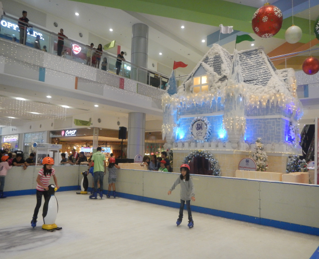 Eine Gruppe von Menschen, die auf einer Eisbahn in einem Einkaufszentrum Schlittschuh laufen, umgeben von einem hausähnlichen Aufbau, der mit Lichtern und festlichen Dekorationen geschmückt ist, mit Zuschauern und zusätzlichen Dekorationen, die eine feierliche Atmosphäre schaffen.