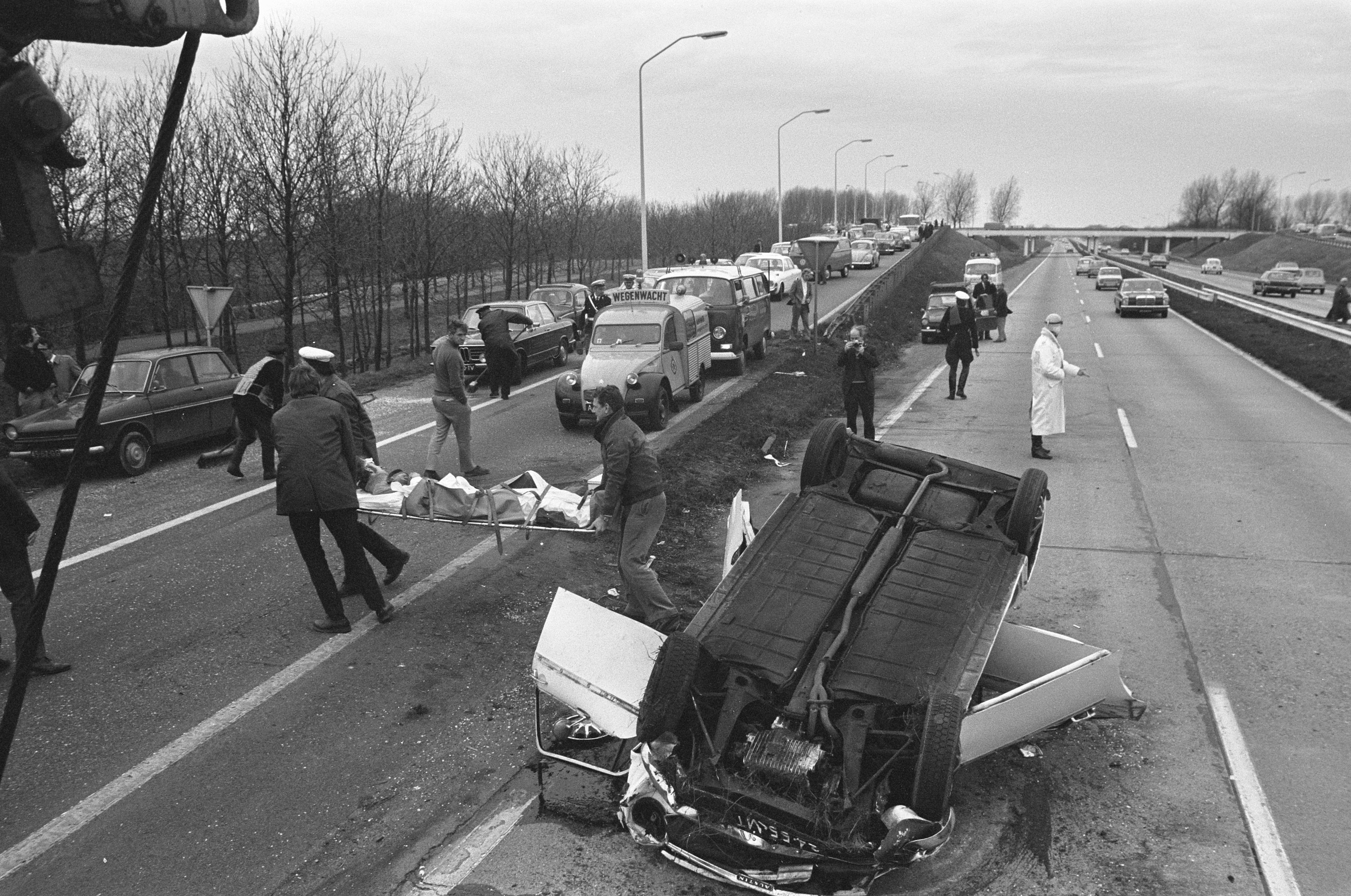 Schwarz-weiß-Foto eines umgestürzten Autos am Straßenrand mit beschädigten Fahrzeugen in der Nähe und einer Gruppe von Menschen drumherum, Lichtmasten, Bäume, eine Brücke und Himmel im Hintergrund.
