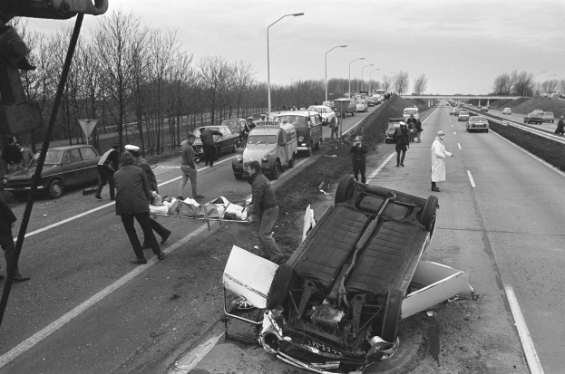 Schwarz-weiß-Foto eines umgestürzten Autos am Straßenrand mit beschädigten Fahrzeugen in der Nähe und einer Gruppe von Menschen drumherum, Lichtmasten, Bäume, eine Brücke und Himmel im Hintergrund.