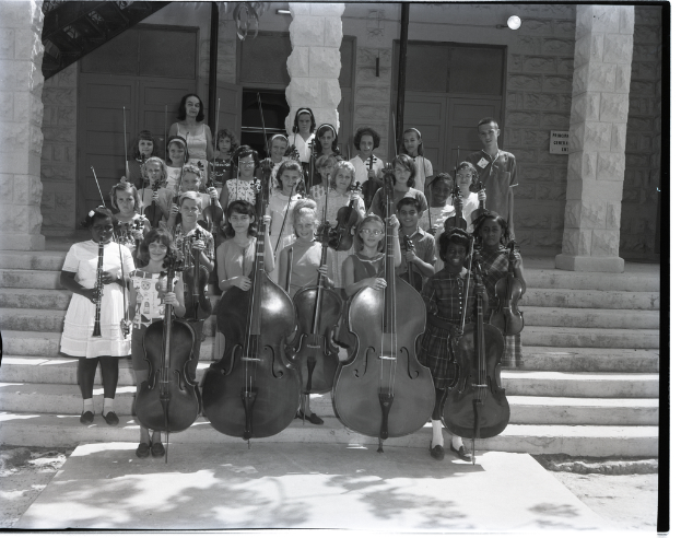 Eine Gruppe von Kindern steht auf einer Treppe und hält verschiedene Musikinstrumente, darunter Celli, Geigen und Trompeten, während im Hintergrund Säulen und eine Wand mit Türen zu sehen sind.