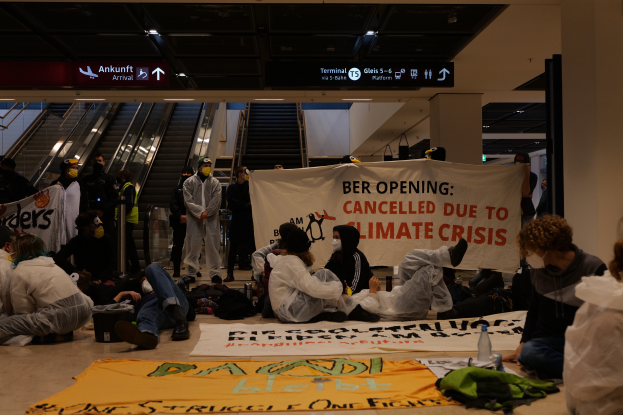 Eine Gruppe von Menschen sitzt auf dem Boden eines Flughafens und hält ein Schild hoch, auf dem "Berlin aufgrund der Klimakrise gestrichen" steht. Es gibt Gepäckstücke, Flaschen und andere Gegenstände um sie herum, und im Hintergrund sind Rolltreppen, Tafeln mit Text und Deckenleuchten zu sehen.