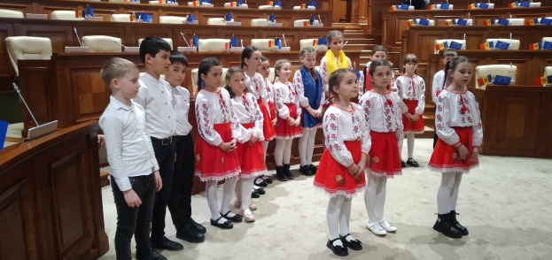 Eine Gruppe von Kindern in weißen und roten Kleidern steht zusammen in einem Raum mit einem Podium und einem Mikrofon auf der linken Seite, Stühle und Tische im Hintergrund und Flaggen, die eine nationale Versammlung anzeigen.