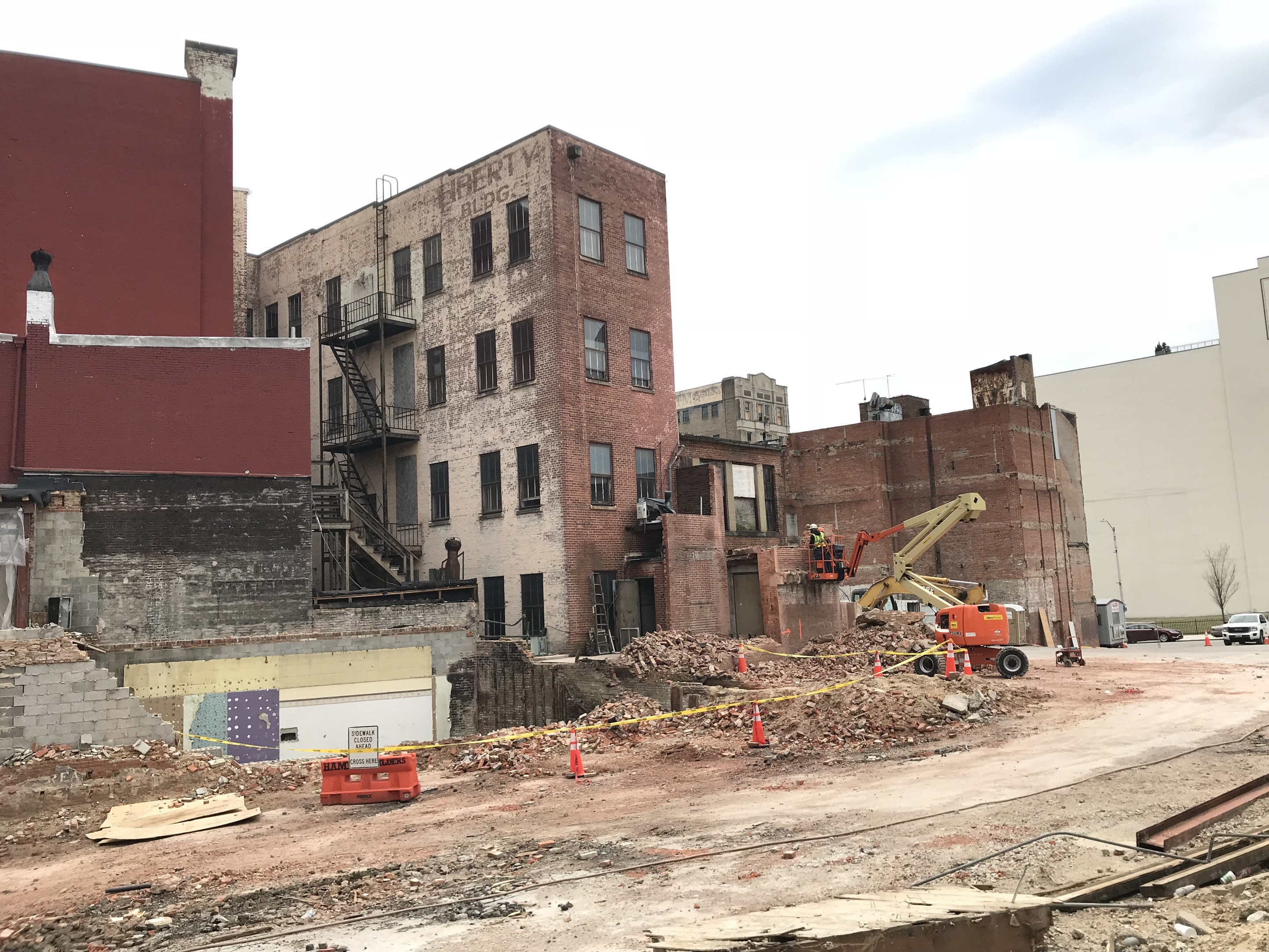 Baustelle mit einem gro├čen Geb├Ąude im Hintergrund, umgeben von Fahrzeugen, Verkehrskegeln, Pfosten, B├Ąumen und Wolken am Himmel.