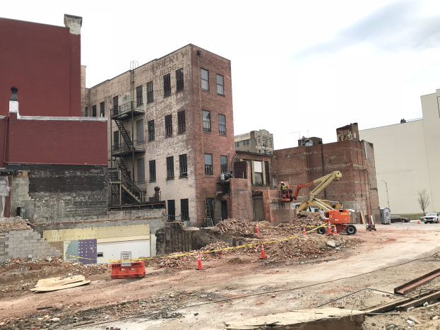Baustelle mit einem gro├čen Geb├Ąude im Hintergrund, umgeben von Fahrzeugen, Verkehrskegeln, Pfosten, B├Ąumen und Wolken am Himmel.