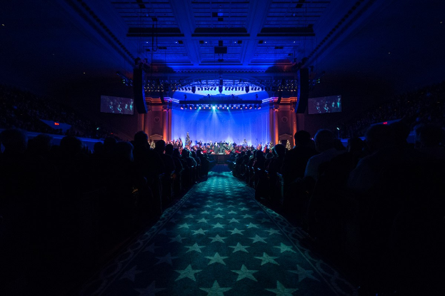 Ein großer Saal mit sitzenden Zuschauern, eine Bühne mit Musikern und Deckenleuchten und -fernseher, was auf eine Konzerthalle für die Nationale Convention 2019 hinweist.