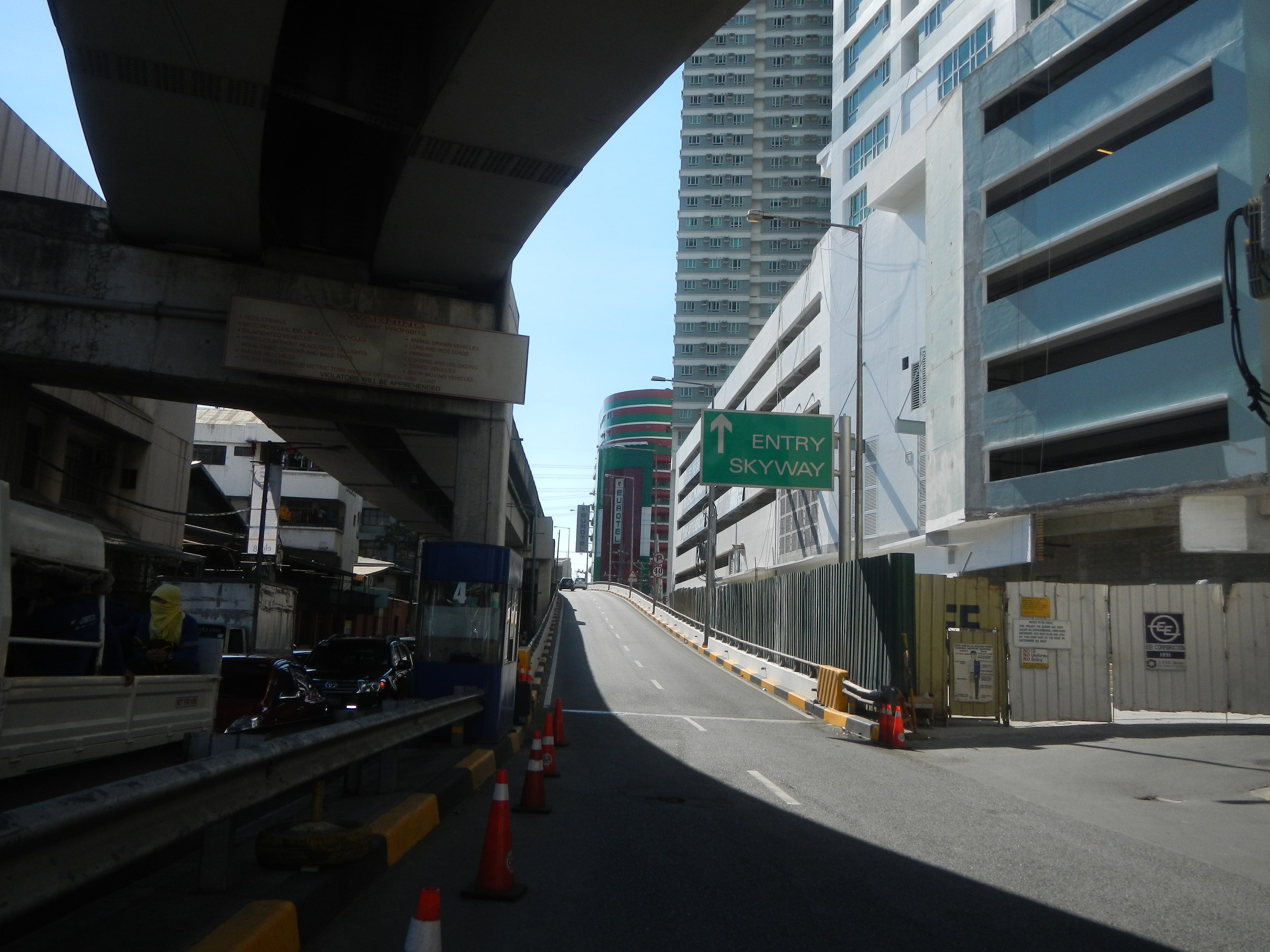 Stadtstraße mit Verkehrskegeln, Fahrzeugen, einer Brücke auf der linken Seite, Gebäuden mit Namensschildern, Laternen und einem Himmel als Hintergrund.