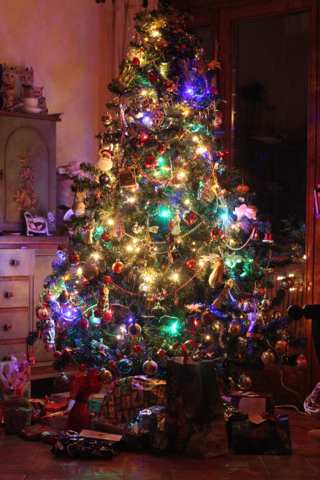 Ein Weihnachtsbaum mit Lichtern und Schmuck, Geschenke auf dem Boden, ein Schrank mit Gegenständen links und ein Fenster auf der rechten Wand.