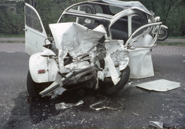 Ein stark beschädigtes Auto steht mit schwerer Frontschaden am Straßenrand, im Hintergrund sind Bäume, ein Gebäude und ein weiteres Fahrzeug zu sehen.