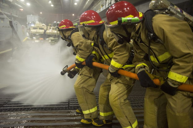 Feuerwehrleute in Helmen und Handschuhen sprühen Wasser auf ein Feuerwehrauto, während sie auf einem Boden mit Hintergrundbeleuchtung, Geländern und Brettern stehen.
