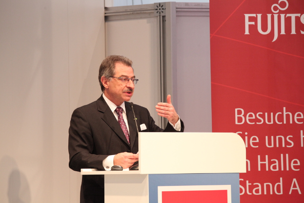 Eine Person mit Brille, Blazer und Krawatte steht neben einem Podium mit Mikrofon und Maus, mit einer Wand und einem Banner im Hintergrund.