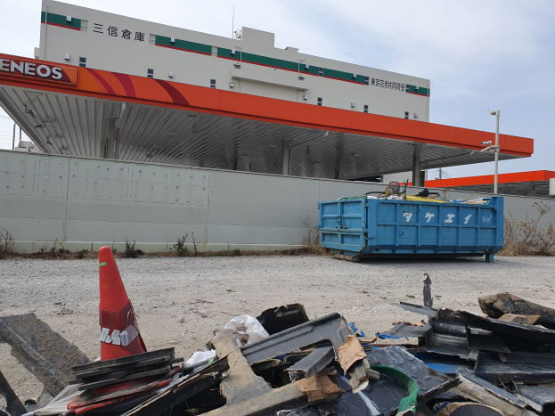 Ein großes Gebäude mit einer beträchtlichen Menge an Schutt, einschließlich Schrottmetall, einem Verkehrskegel und einem blauen Container davor, sowie Pflanzen, Polen und Himmel im Hintergrund, was auf Abrissarbeiten hinweist.