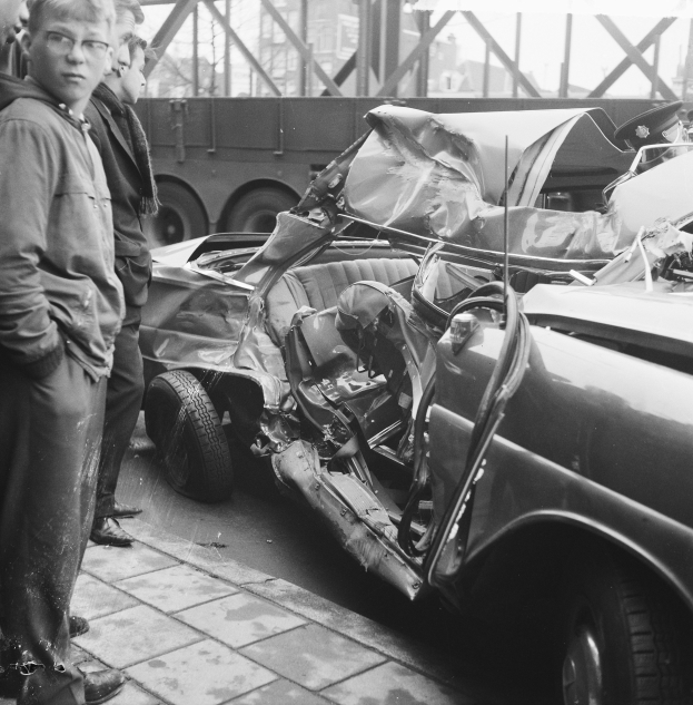 Ein Schwarz-Weiß-Bild eines verunglückten Autos am Straßenrand mit mehreren Personen drumherum, einem Lastwagen und einer Brücke im Hintergrund.