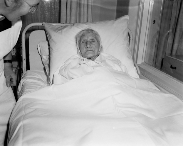 Ein älteres Bild einer Frau im Krankenhausbett mit einem weißen Laken und Kissen, während eine Krankenschwester daneben steht, mit einem Fenster mit Vorhängen im Hintergrund.