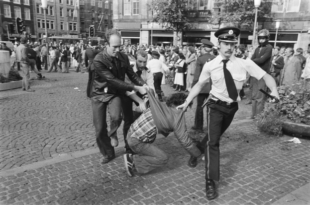 Schwarz-weißes Bild eines Mannes, der von zwei Polizisten auf einer Straße mit Zuschauern, Gebäuden, Bäumen und Ampeln im Hintergrund gezogen wird.