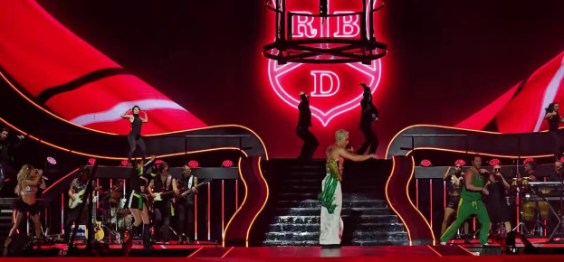 Eine Gruppe von Menschen auf einer Bühne, einige halten Musikinstrumente, mit Stufen, die zur Bühne führen und einem Schild im Hintergrund mit der Aufschrift "Rihanna - Live im O2 Arena, London".