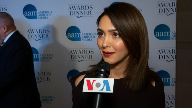 Emmy Rossum in einem schwarzen Kleid hält ein Mikrofon, während ein Mann links neben ihr steht und ein Banner mit der Aufschrift "Aam Awards Dinner" im Hintergrund zu sehen ist.