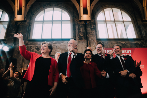 Eine Gruppe von Menschen, wahrscheinlich Labour-Parteimitglieder, steht zusammen klappend in Feierlaune vor einer Menge, mit einem Banner und beleuchteten Fenstern im Hintergrund.