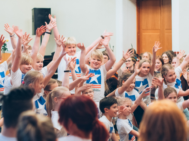 Eine Gruppe von Kindern hebt die Hände vor einer Menge, mit einer Wand und einer Tür im Hintergrund, in der Nähe eines Tisches mit einer Blumenvase und einem Lautsprecher.