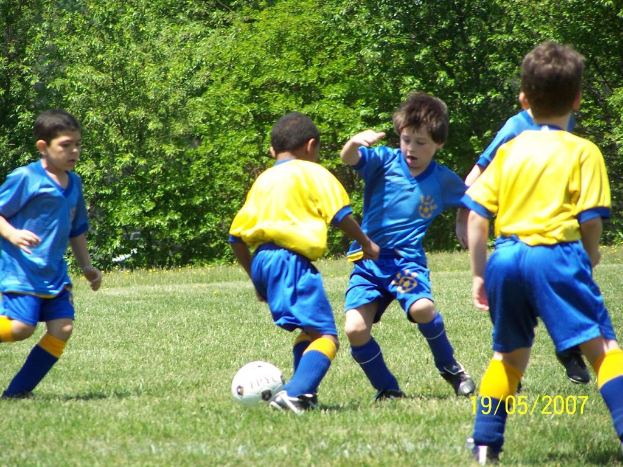 Kinder beim Fußballspielen auf einer grünen Wiese mit Bäumen im Hintergrund und einem Datum in der rechten unteren Ecke.