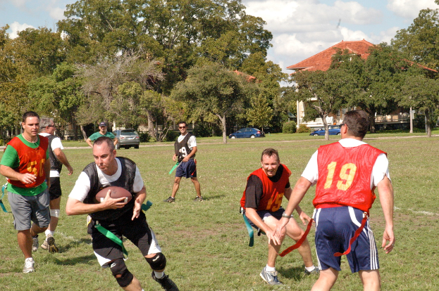 Leute, die American Football auf einem grasbewachsenen Feld mit einem Haus, Autos und Bäumen im Hintergrund spielen.