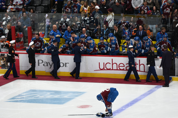 Ein Hockey-Spieler auf dem Eis mit einem Stock in der Hand, umgeben von einer Gruppe von Menschen mit Mützen und Hockey-Stöcken, mit Zuschauern und Brettern, die "Colorado Avalanche vs San Jose Sharks - 3/24/15 NHL Pick, Odds, and Prediction" anzeigen.