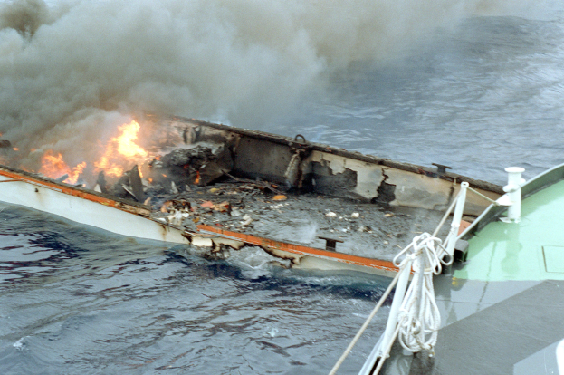 Ein brennendes Boot auf dem Meer mit dicken Rauchschwaden und sichtbaren Tauen auf der rechten Seite.