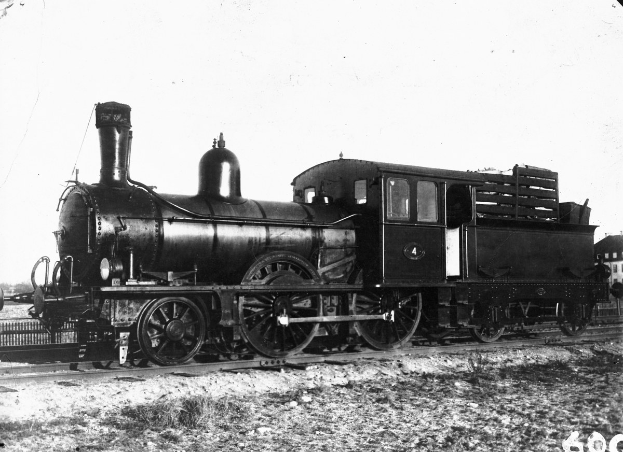 Schwarz-weißes Foto einer Dampflokomotive auf einem Bahngleis, umgeben von einem Zaun, Gebäuden, Bäumen und einem klaren Himmel, mit Text unten.