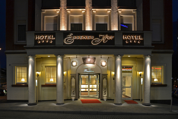 Das Essener Hof Hotel in Berlin, Deutschland, ein mehrstöckiges Gebäude mit Fenstern, Türen, Säulen und Lichtern, mit einem Namensschild sichtbar, und einer Straße mit Fahrzeugen auf der rechten Seite, unter einem klaren Himmel.