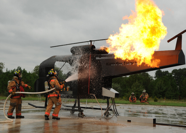 Feuerwehrleute spritzen Wasser auf einen brennenden Hubschrauber in einem waldigen Gebiet unter einem klaren blauen Himmel.