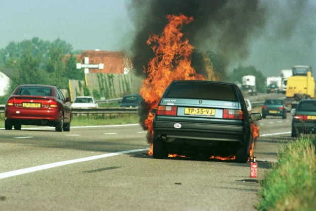 Ein Auto in Flammen am Straßenrand mit umliegenden Fahrzeugen, Bäumen, Gebäuden und einem klaren blauen Himmel im Hintergrund und einem Feuerlöscher auf dem Gras auf der rechten Seite.