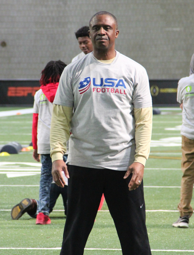 Ein Fußballer in Stutzen steht auf einem Feld mit einer Gruppe von Menschen und einer Wand im Hintergrund.